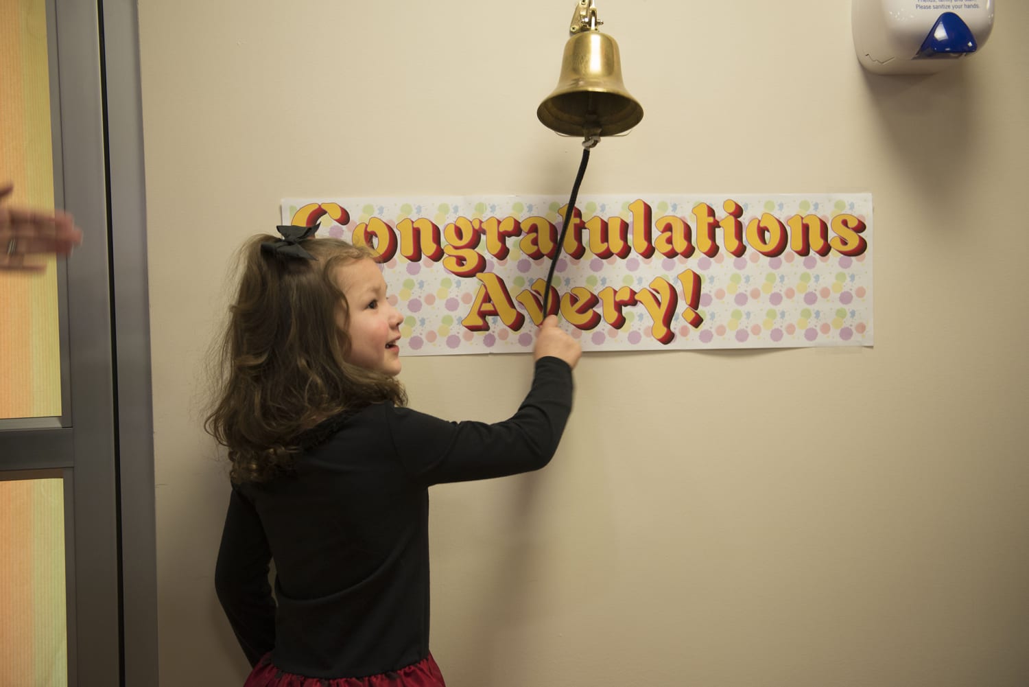 Beige wall with a bell and underneath a banner saying Congratulations Avery! Little girl with a bow in her hair, ringing the bell