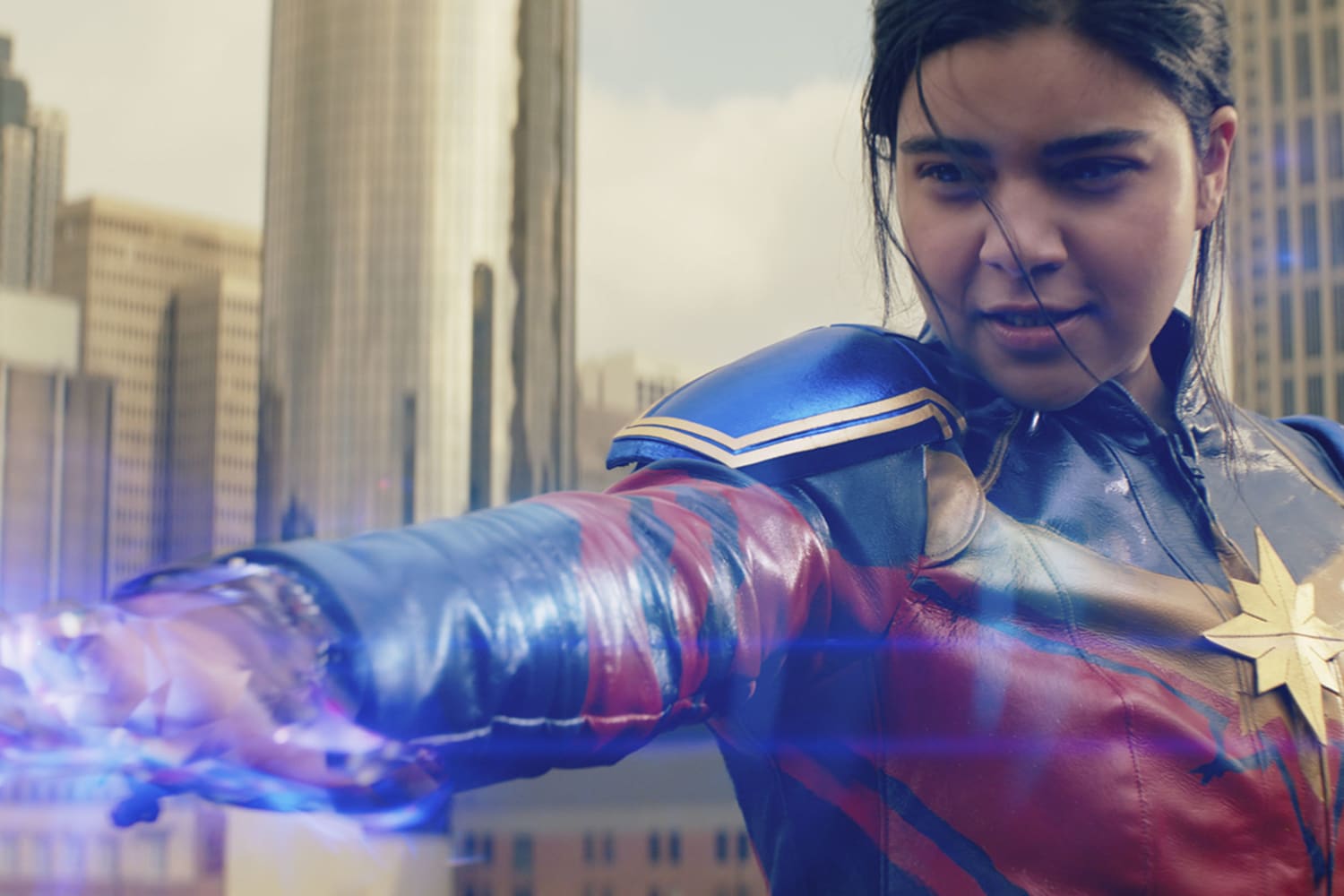 Young Pakistani woman looks at her own outstretched arm. Her hand is open and lined with white, blue and purple light. She is wearing a navy blue, red and gold leather costume with a star on her chest. The background is a blurred city skyline