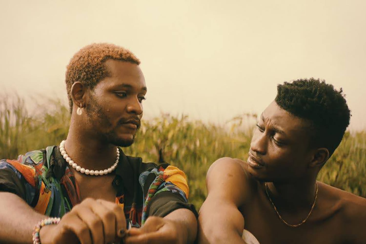 Two Black men sitting in the grass, exchanging a tender glance