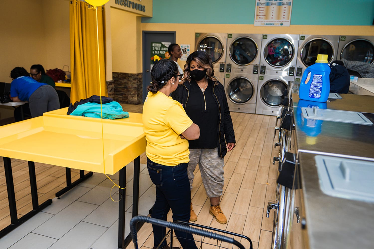 Two women wearing masks in conversation in a laundromat. They stand next to a large yellow table with a yellow balloon, in front of a row of laundry machines. Two women are speaking at a table to the left of them, and one stands behind them.