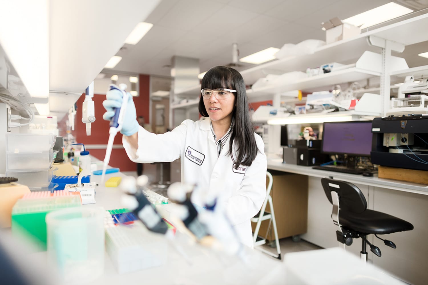 A woman with long dark hair and bangs stands in a lab holding a syringe out in front of her, pouring liquid into tubes on a lab table covered with charts and other tubes. Behind her, a desktop computer, a computer chair, and shelves with paper and folders