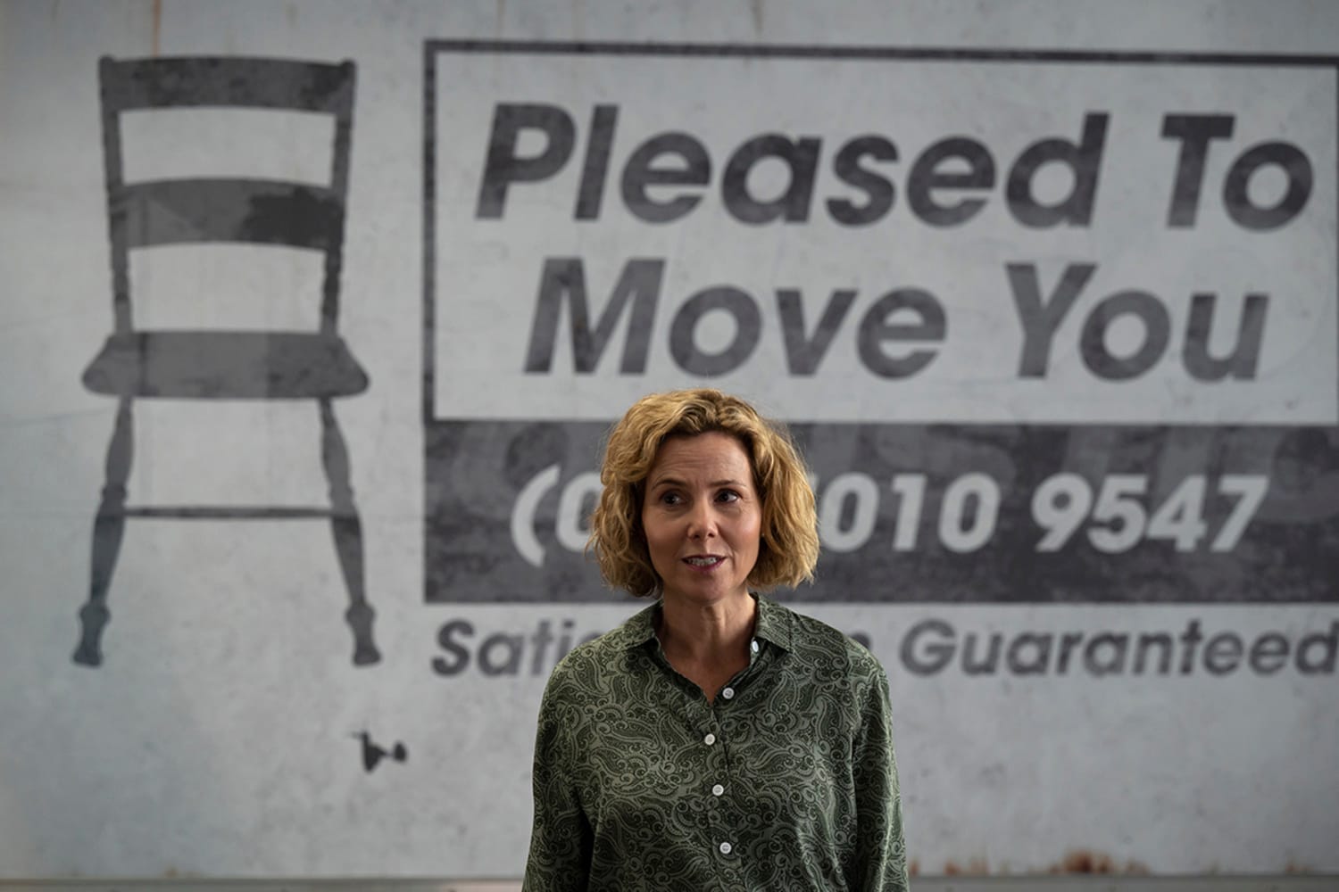 A woman with short, curly blonde hair stands speaking in front of a large business sign that reads: Pleased to Move You (partially covered phone number, "Satisfaction guaranteed" printed below). An image of a chair appears to the left of the business sign