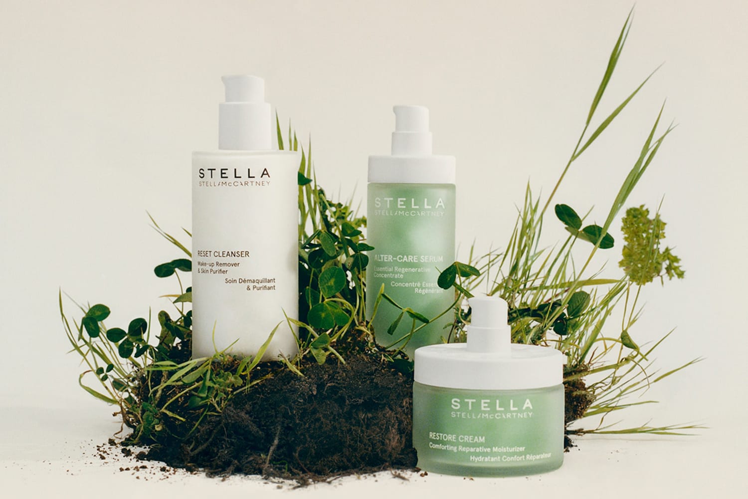 Three bottled products situated around a grassy, uprooted lump of dirt. Left to right: Stella Reset Cleanser, Stella Alter-Care Serum, Stella Restore Cream.