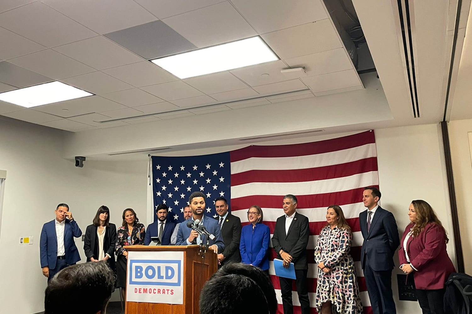 A dark-skinned man wearing a navy suit stands at a podium speaking into several microphones. A sign on the podium reads: BOLD Democrats. He stands in front of eleven adults and a large American flag.