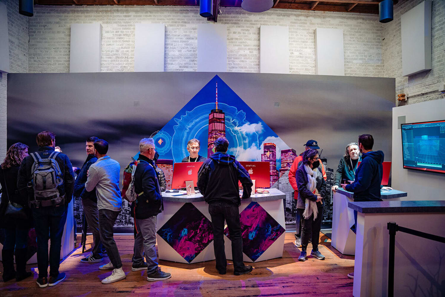 Several people interacting in a small room with a table in the center and two desktop screens. A blue and red backdrop of the Freedom Tower rests behind them.