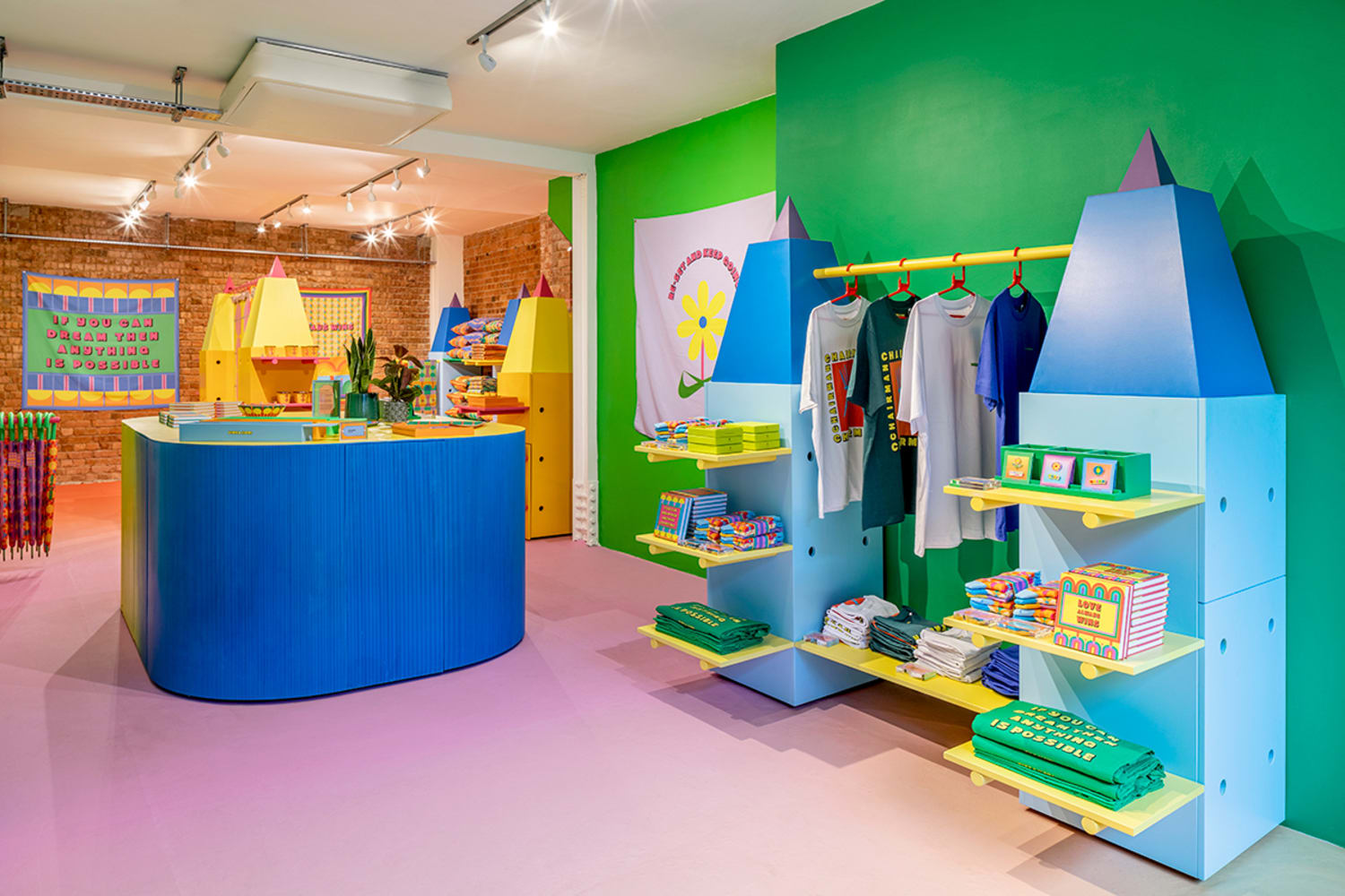 A colorful storefront with pink flooring, a green wall to the right, and a blue counter to the left with tapestries, clothing and books covering the walls and shelves.