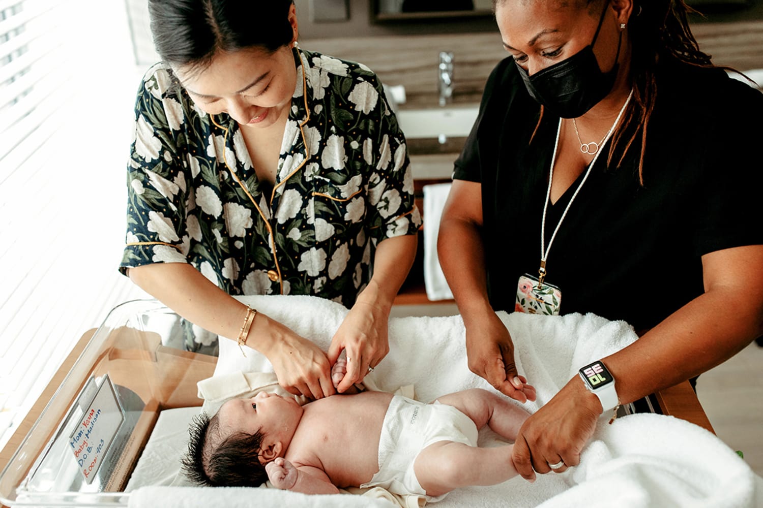 Two women stand over a baby in a bassinet. The woman on the left has light skin and black hair and wears a floral button-down pajama top. On the right, a dark-skinned woman with braids wears a black mask and a black shirt.