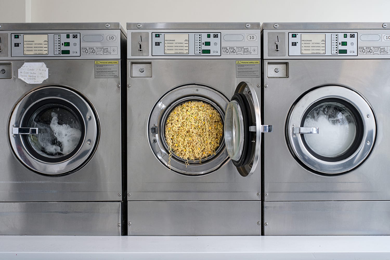 Three large industrial washing machines, silver and chrome colored, with front-facing doors and a window. In the middle washer, the door is open and noodles overflow out of the open door.