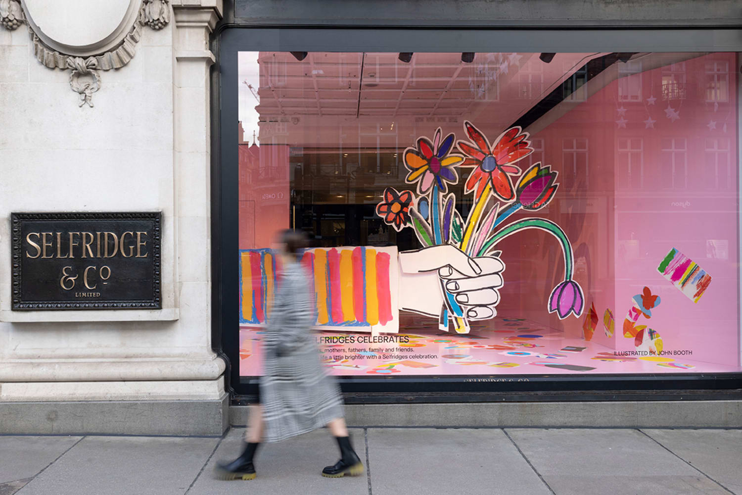 A woman blurred by movement walks by a pink window display of a giant cartoonish hand holding colorful giant cartoonish flowers. To the left, a sign reads Selfridge & Co.