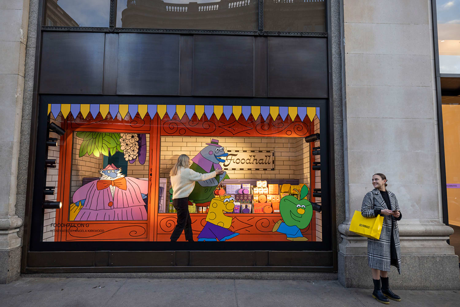 A woman with blonde hair is blurred in movement as she stands inside a colorful window display adjusting its contents: a cartoonish character with blue skin and an orange suit serves other cartoon characters. Another woman stands just outside the window.