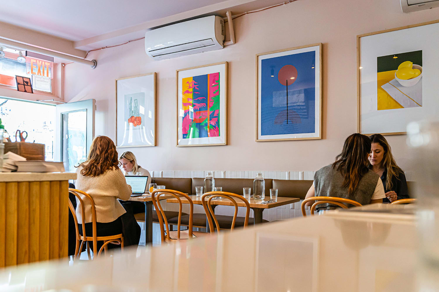 A set of 4 small tables, with two people sitting at the farthest table on the left and one person sitting at the farthest on the right, in front of four colorful paintings hung on the wall above a booth.