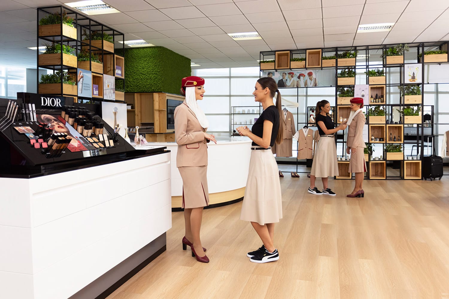 Two women stand facing each other in a beauty retail space. On the left, a woman wearing a tan flight crew uniform and a red cap. On the right, a woman with dark hair wears a white long skirt and a black short sleeve shirt.