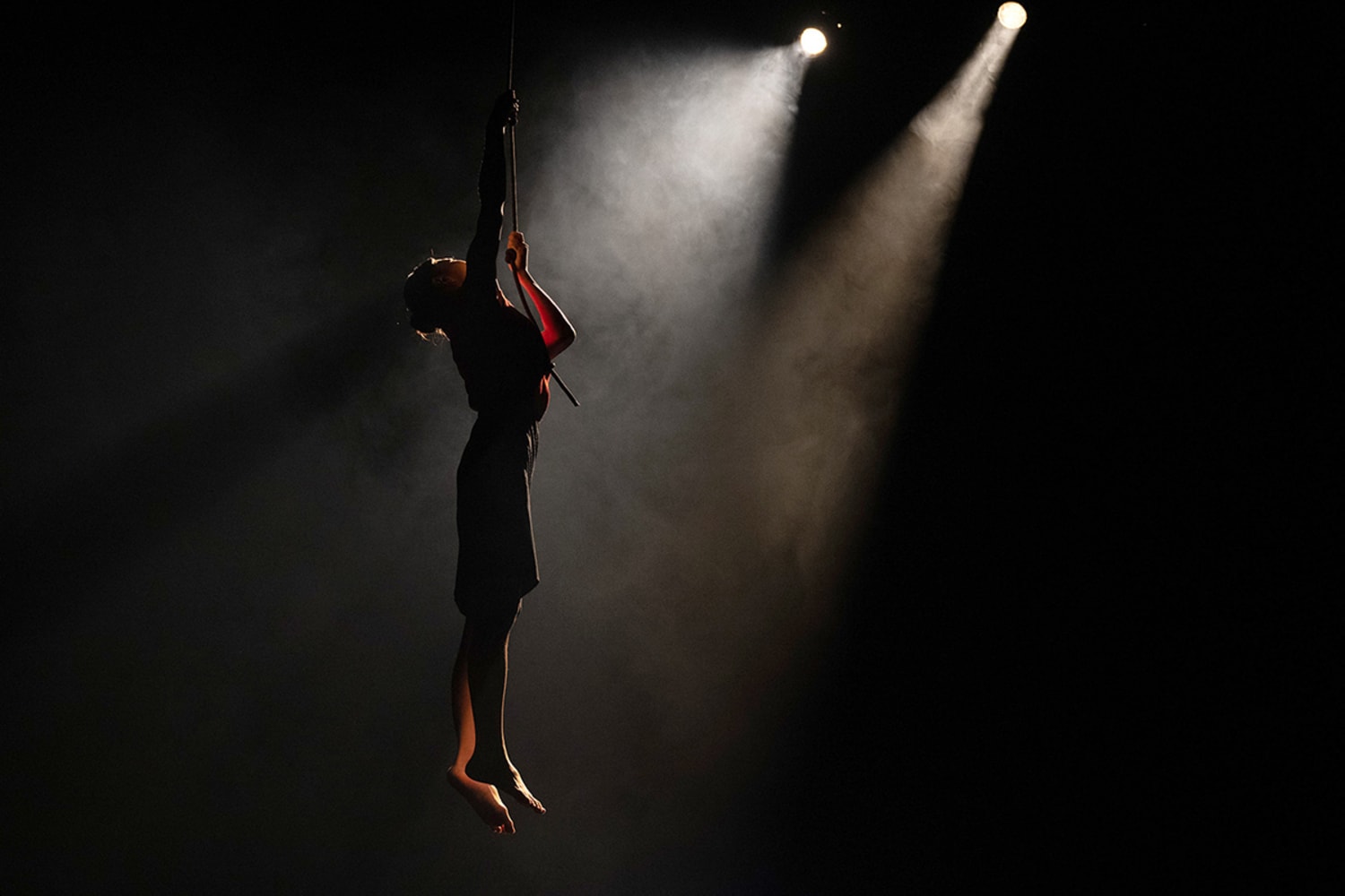 A woman climbs up a rope, her legs straight below her, with darkness and a small spotlight behind her.