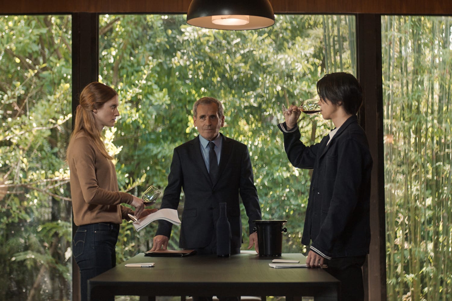 A man with dark hair and a woman with light red hair stand across from each other at a small square table. An older man stands between them, facing the viewer. The man, on the right, sips wine from a glass.