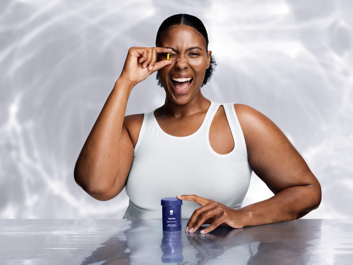 Black woman wearing a white formfitting vest with raindrop cutout at shoulder She is sat at a table with shiny surface and holding a yellow capsule in front of her right eye and smiling