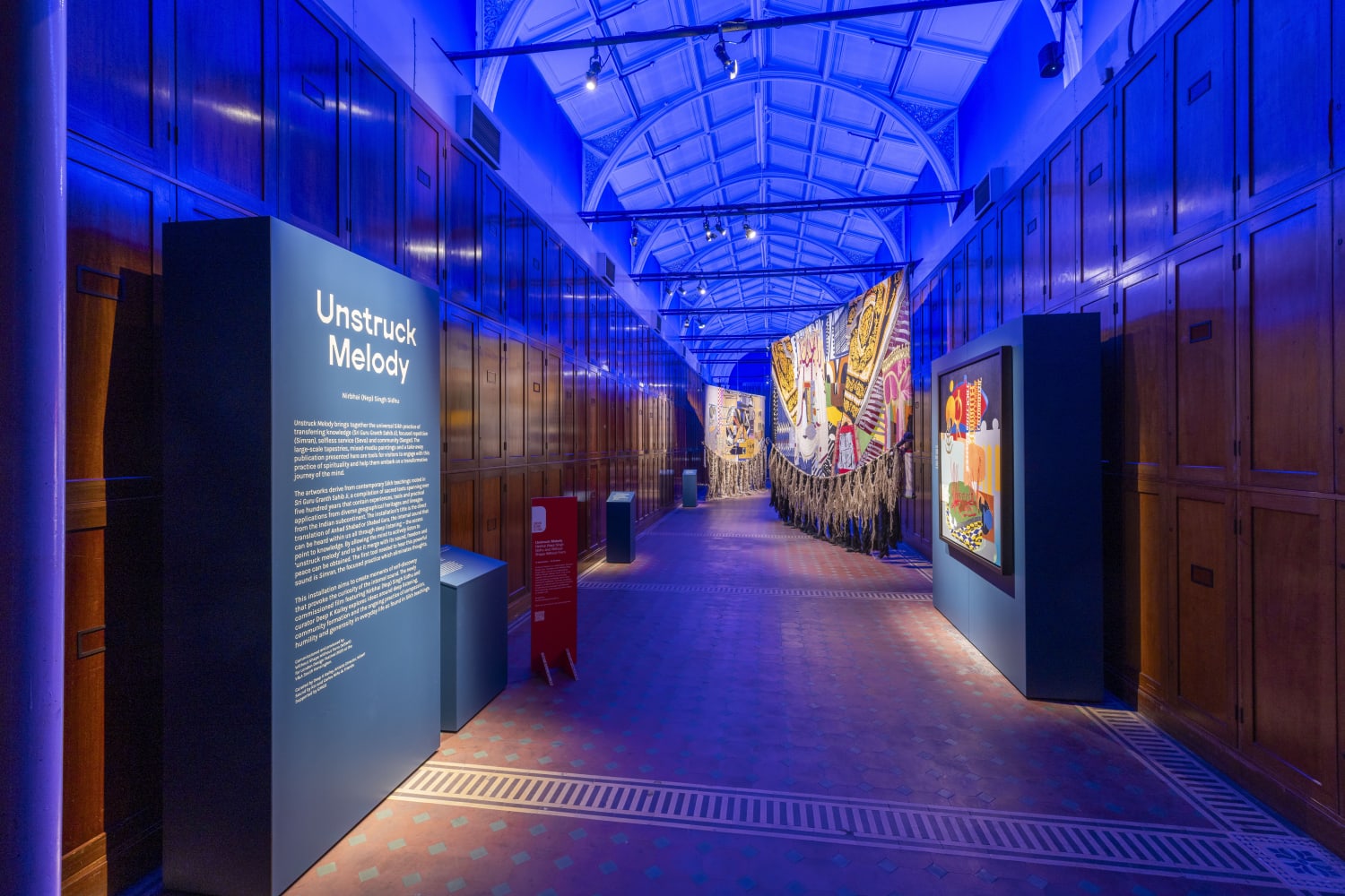 Long arched room bathed in blue light in which large tapestries are displayed with an information board at the front with the words Unstruck Melody