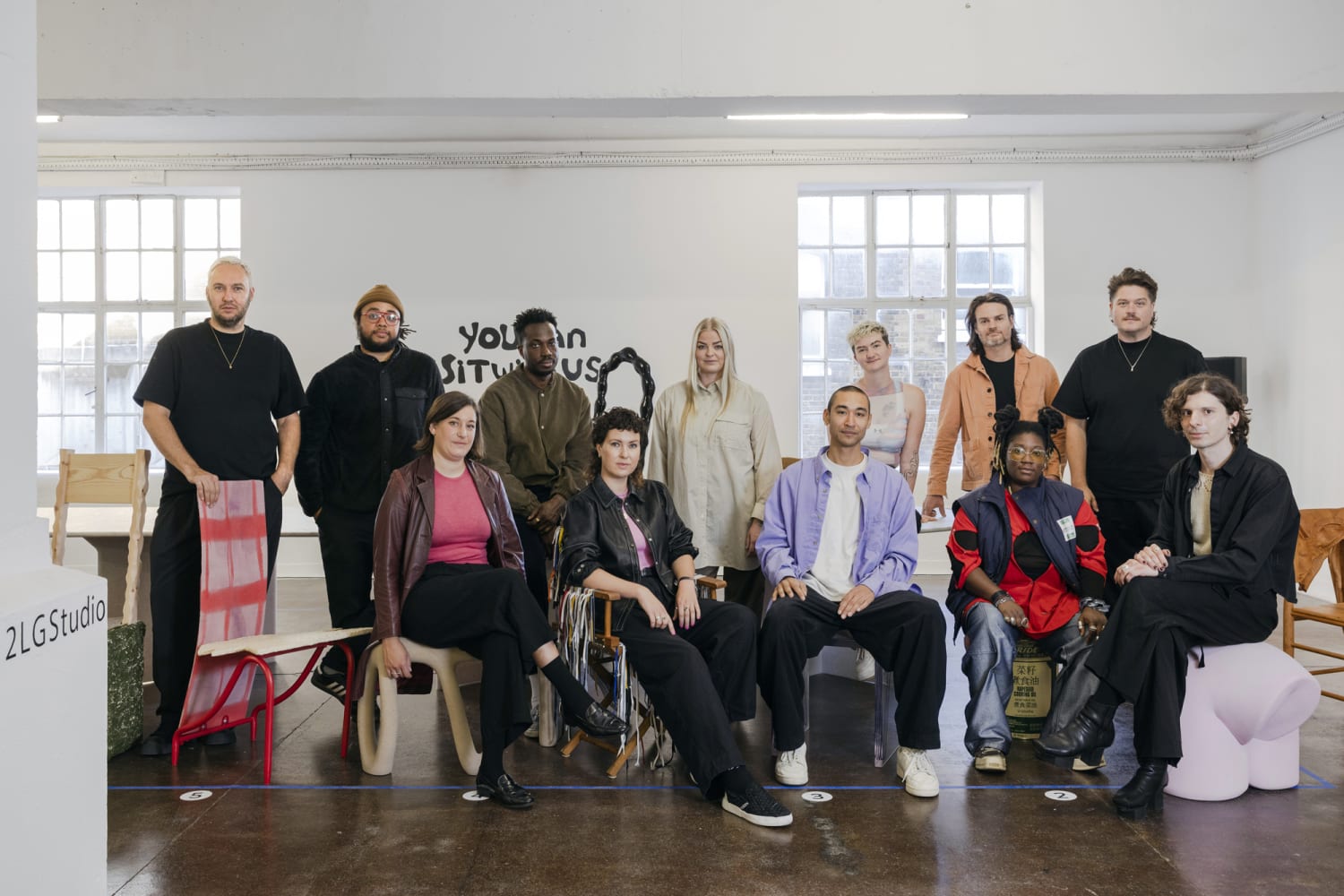 Diverse mixed group of 12 people sitting in a studio some seated some standing Behind them on the wall is text You Can Sit With Us