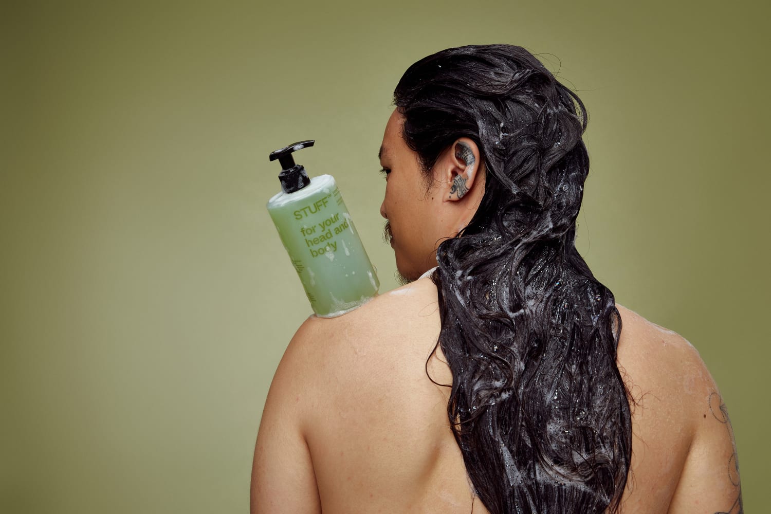 A male with long wet dark hair has his back to the camera with a bottle branded Stuff on his shoulder