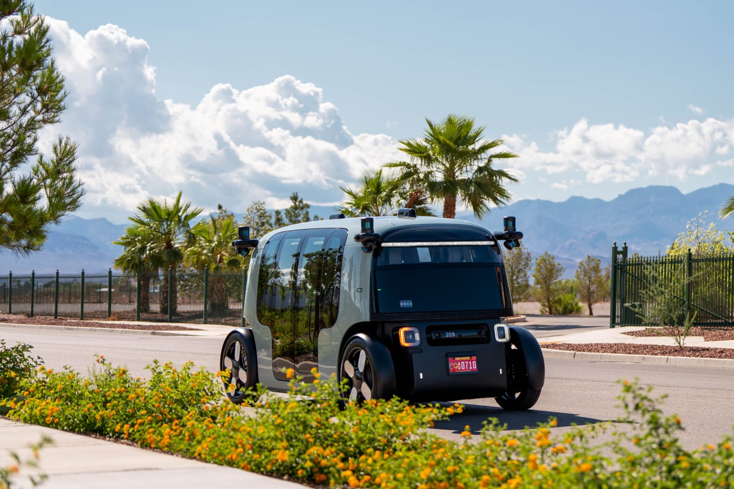 A sage green and black robotaxi shuttle is parked on a Las Vegas street In the background are palm trees and mountains