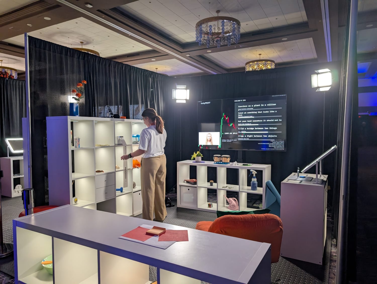 An installation of a sqaure space room surrounded by shelves and objects with a large screen on which instructions for tasks are appearing. A women in grey top and green slacks is performing a task.
