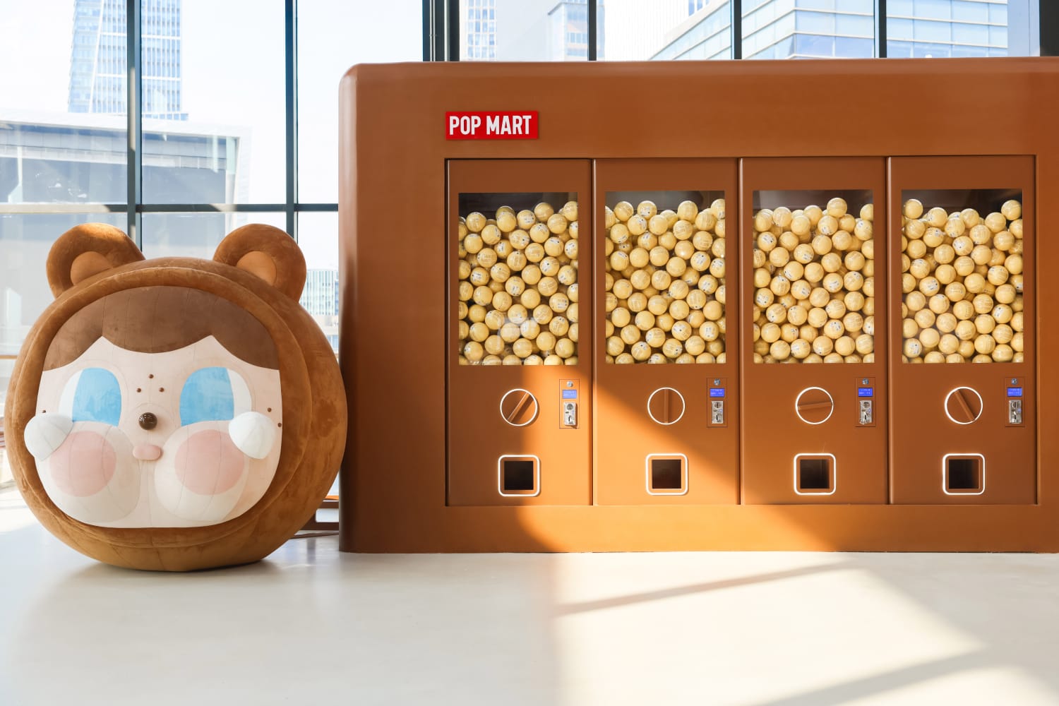 Giant brown plushy bear-face beside a POP MART display case filled with small boxed figures in front of windows.
