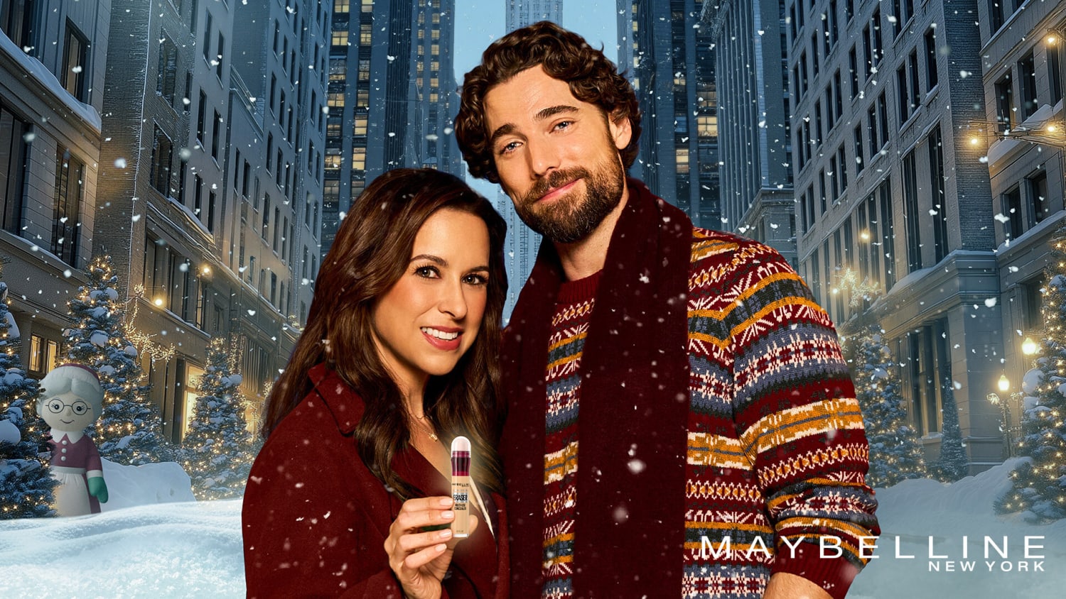 A dark-haired couple stand in the snow smiling, against a New York backdrop. The woman is holding a Maybelline foundation bottle.