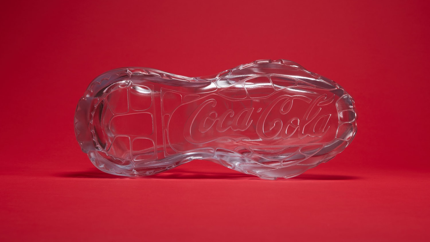 A bottom view of a transparent glass sculpture shaped like a sneaker, displayed against a solid red background.
