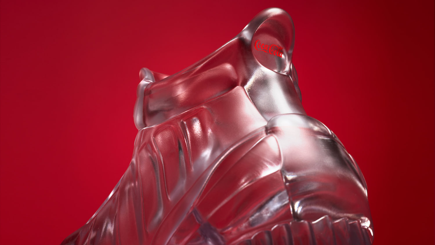 A back heel view of a transparent glass sculpture shaped like a sneaker, displayed against a solid red background.