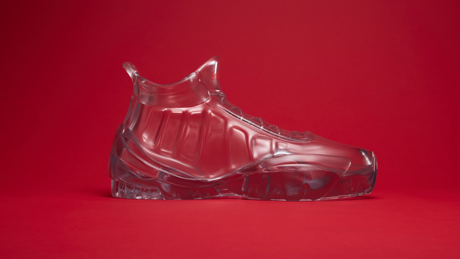 A side view of a transparent glass sculpture shaped like a sneaker, displayed against a solid red background.