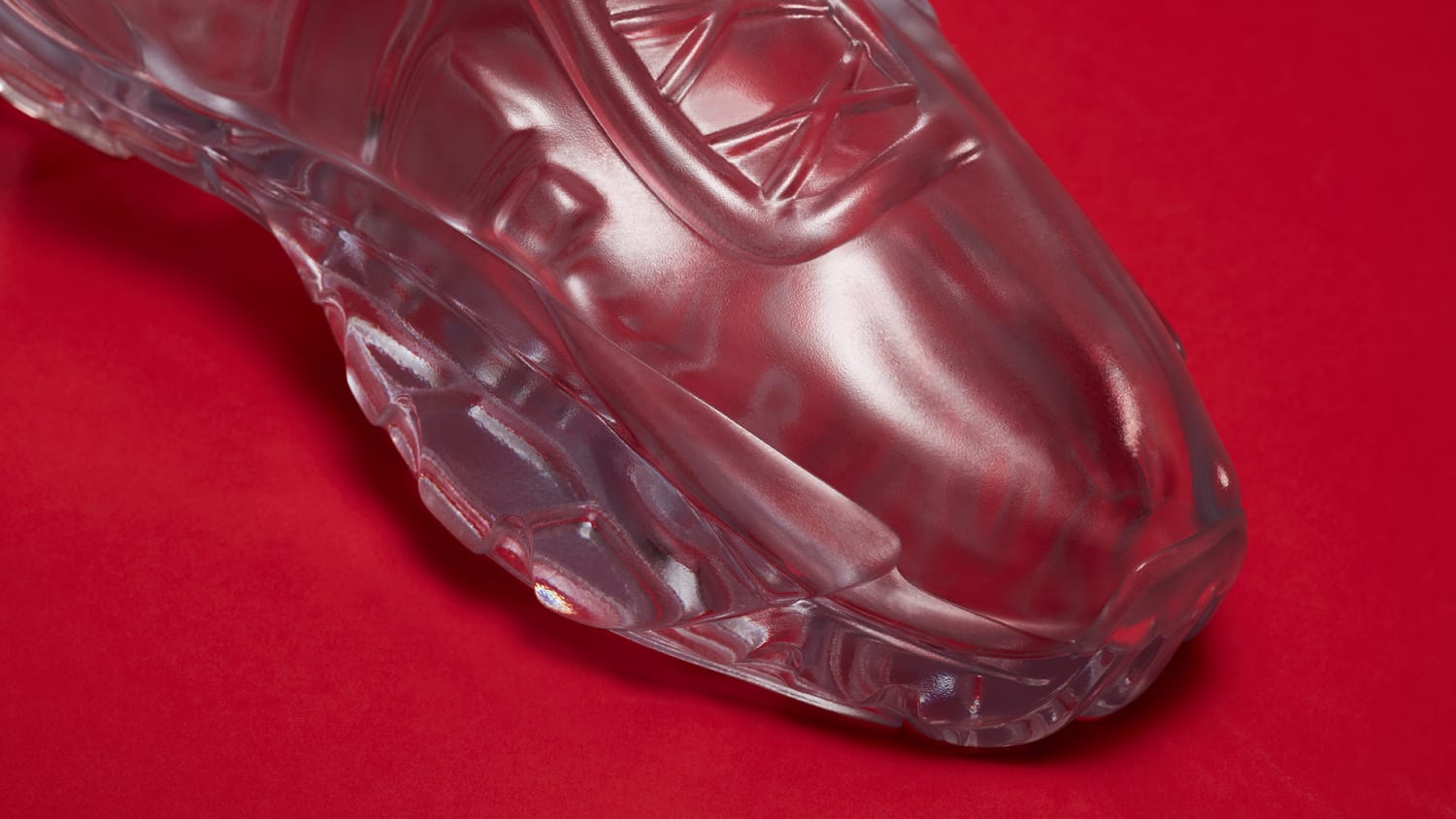 A toe view of a transparent glass sculpture shaped like a sneaker, displayed against a solid red background.