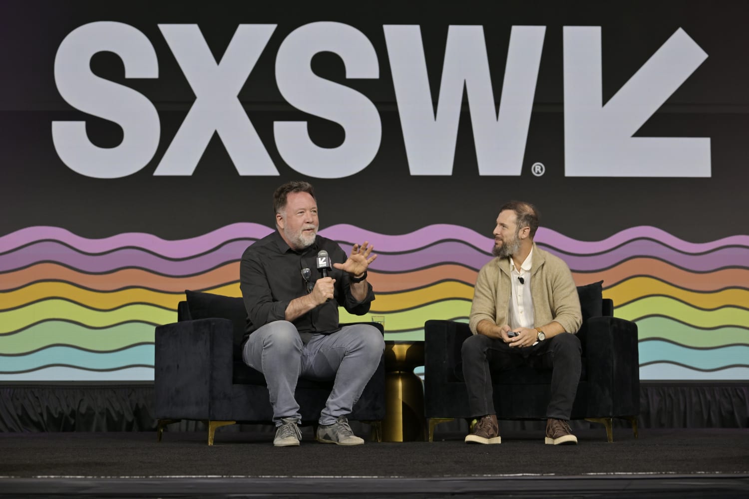 Two men - Aza Raskin and Steven VanRoekel - have a fireside chat at SXSW 2026. They are dressed casually and sitting in armchairs beneath a SXSW logo banner.