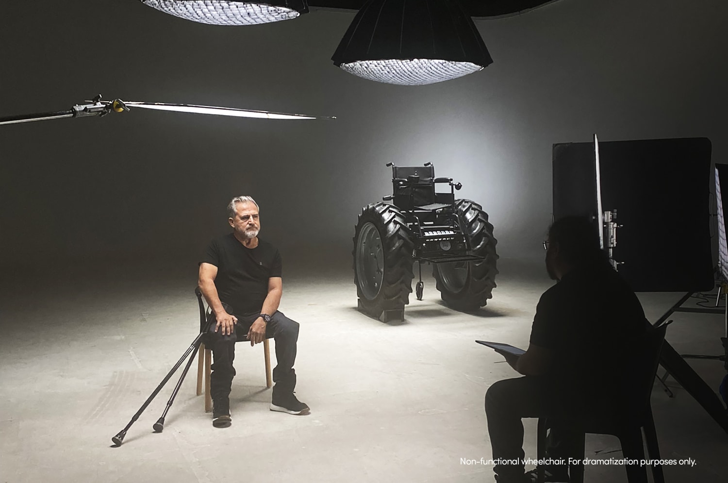 En un estudio iluminado, el hombre protagonista de la campaña 'SILLA MONSTER' se sienta mirando fijamente la imponente silla de ruedas con ruedas de tractor, mientras una persona del equipo técnico trabaja en primer plano. Esta imagen captura el detrás de escena de la visibilización de la problemática de la accesibilidad en Argentina.