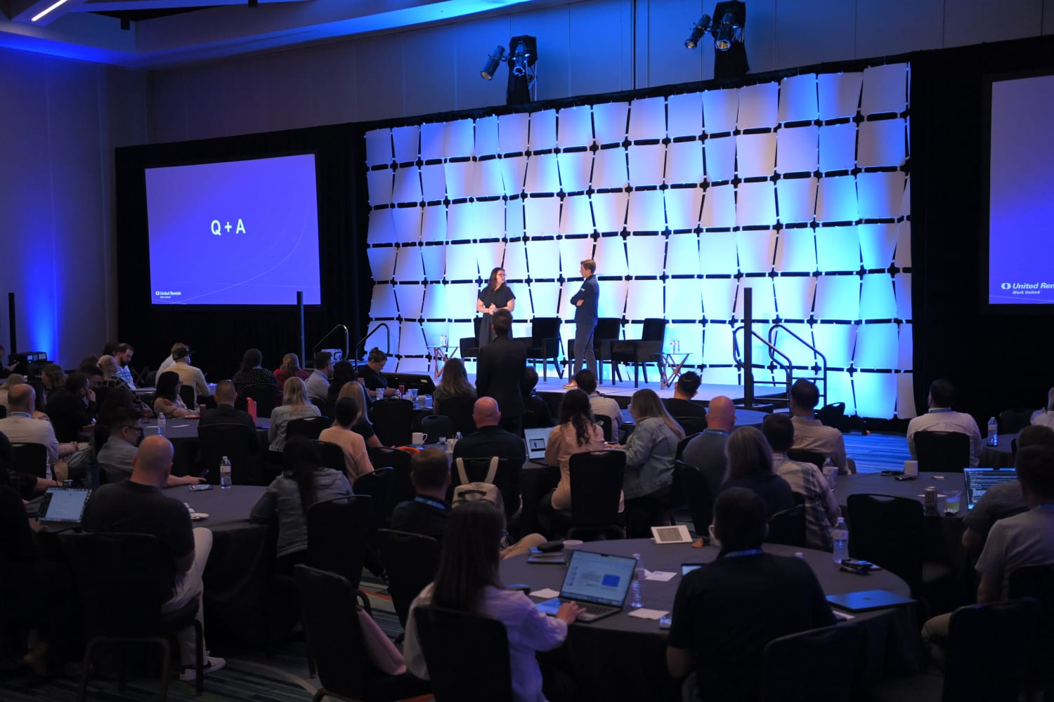 Two people standing on stage at an indoor event in front of a lively and full audience.