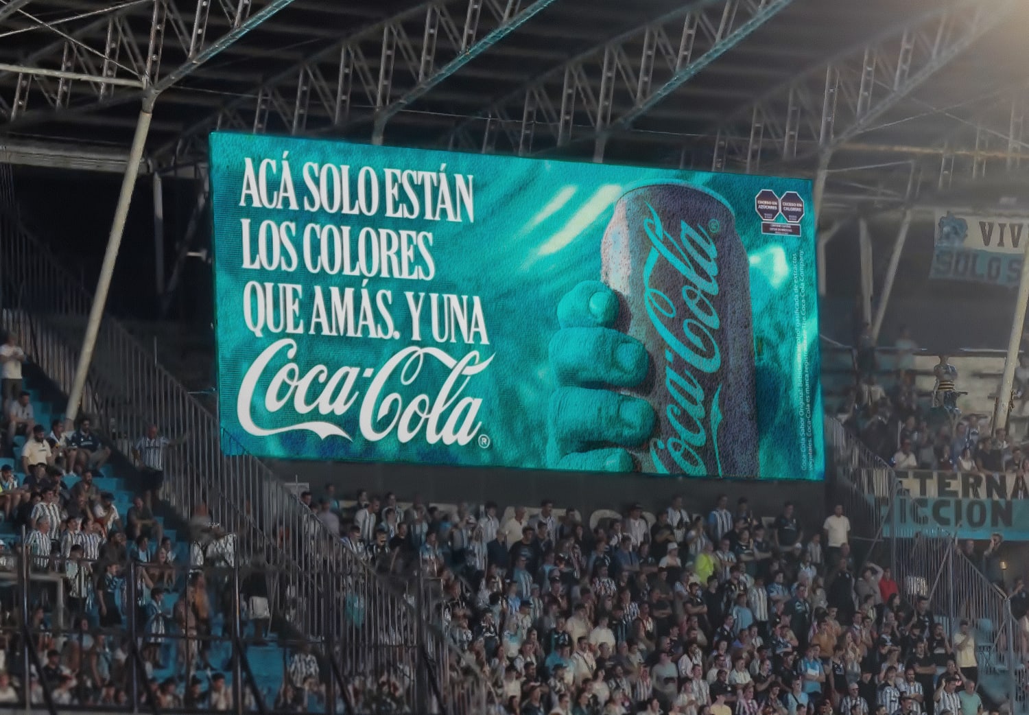 A large crowd is gathered in a stadium. The focus is on a prominent Coca-Cola advertisement displayed on a digital screen, featuring a hand holding a bottle. The ad text is in Spanish.