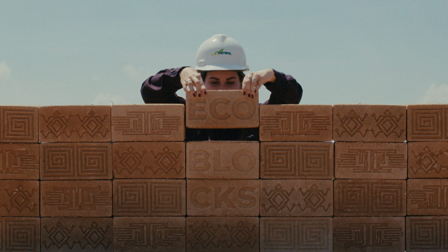 A worker in an Ecopetrol hard hat peers over a wall of patterned "ECO BLOCKS" against a clear sky.