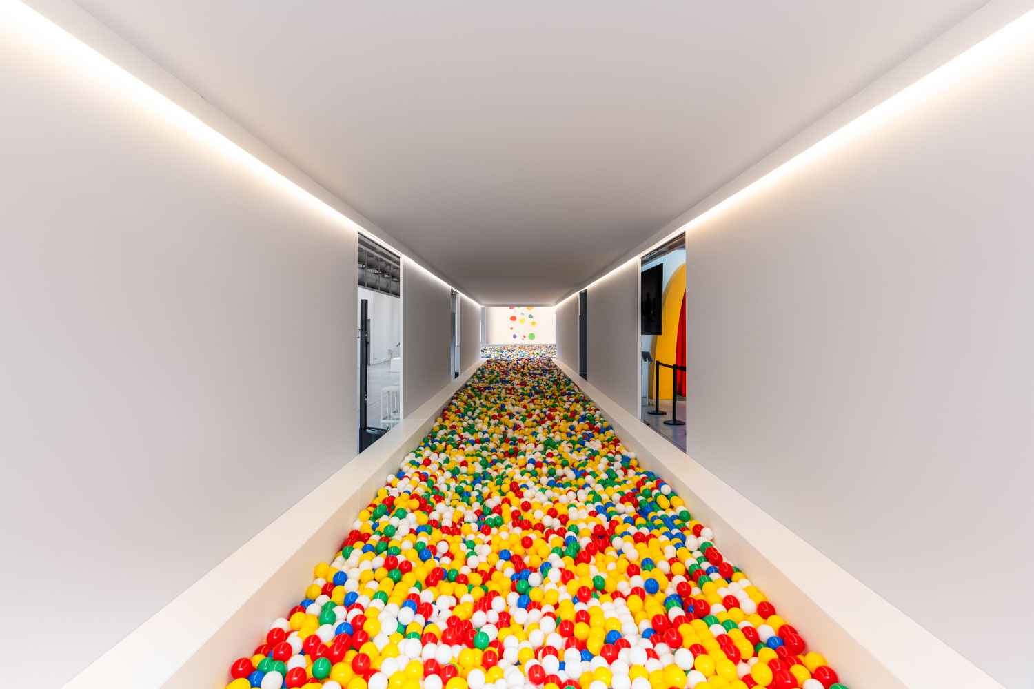 A long, sleek, white hallway stretches into the distance, with its entire floor transformed into a vibrant ball pit. The pit is overflowing with countless red, yellow, green, blue, and white plastic balls. Recessed lighting illuminates the clean, minimalist walls and ceiling, creating a playful yet modern aesthetic.