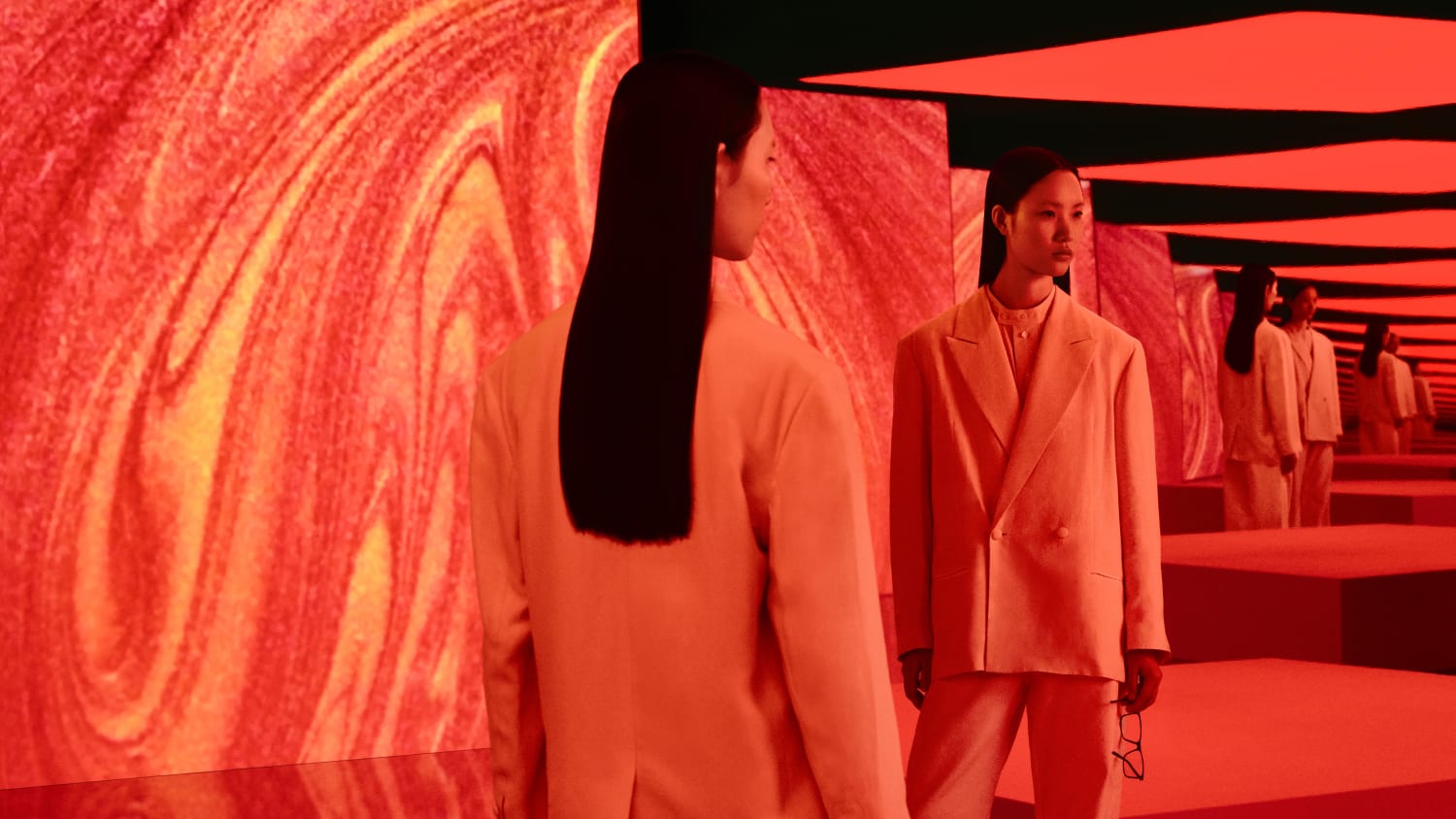 Two women in light-colored suits stand in an immersive room with walls covered in vibrant red, orange, and yellow swirling, molten-like patterns. Reflections of the women and the patterned walls extend into the distance, creating a sense of depth