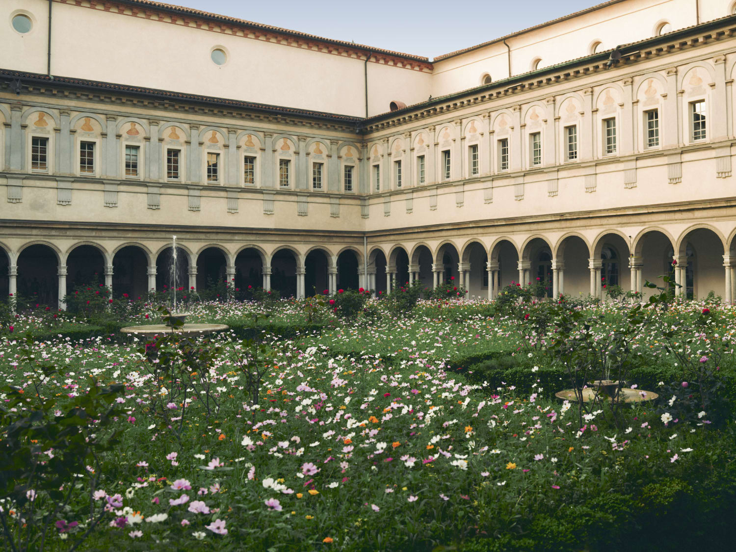 A classical architectural courtyard featuring a lush garden filled with numerous light-colored flowers and green foliage. The garden is surrounded by a multi-story building with an arched colonnade on the ground floor and rows of windows on the upper levels.