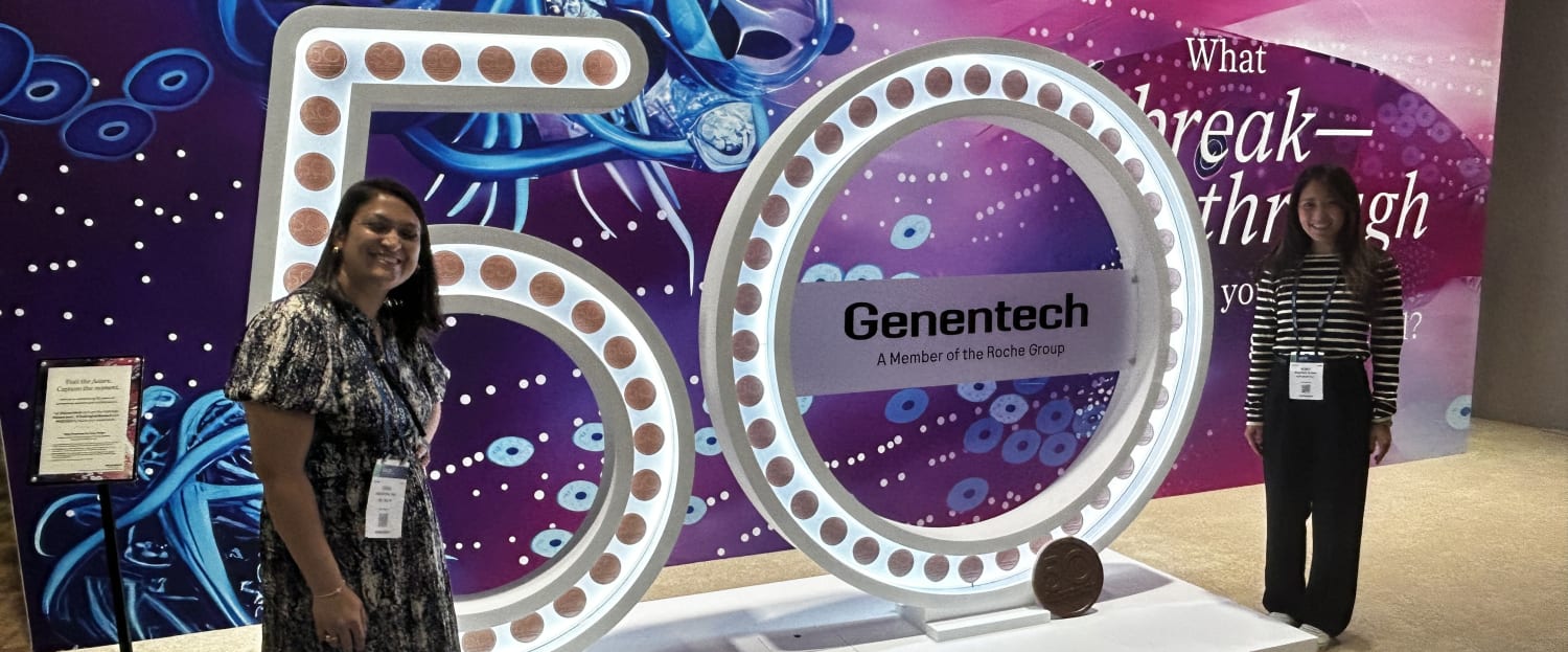 Two women smile next to a giant illuminated '50' sign featuring the Genentech logo, set against an abstract purple and pink cellular background.