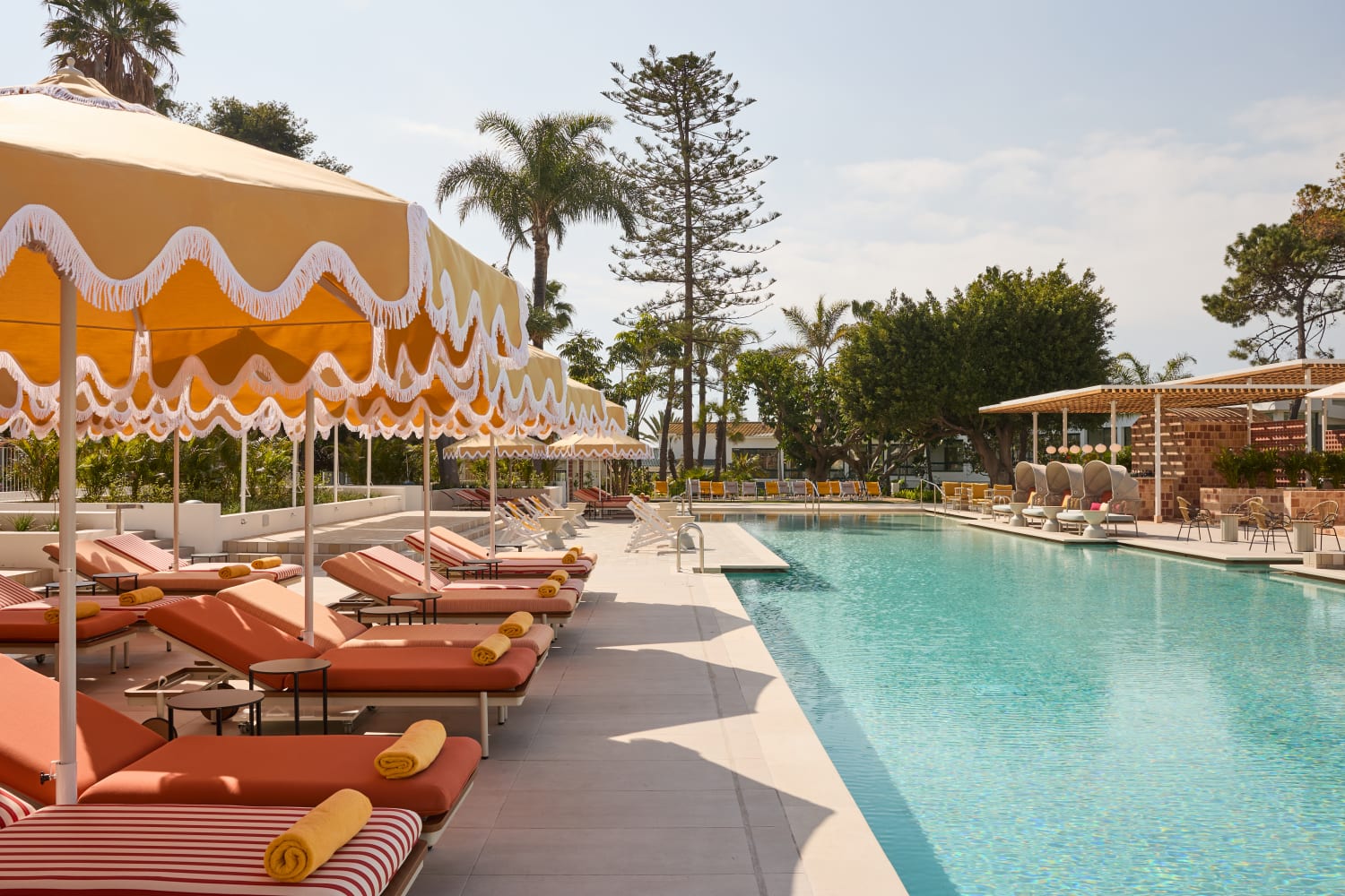 A sunny, luxurious outdoor resort pool area featuring a long, clear blue swimming pool. Alongside the pool are rows of orange-and-white striped lounge chairs shaded by retro-style yellow umbrellas with white scalloped trim. Lush green palm trees and foliage are visible in the background under a bright, clear sky.