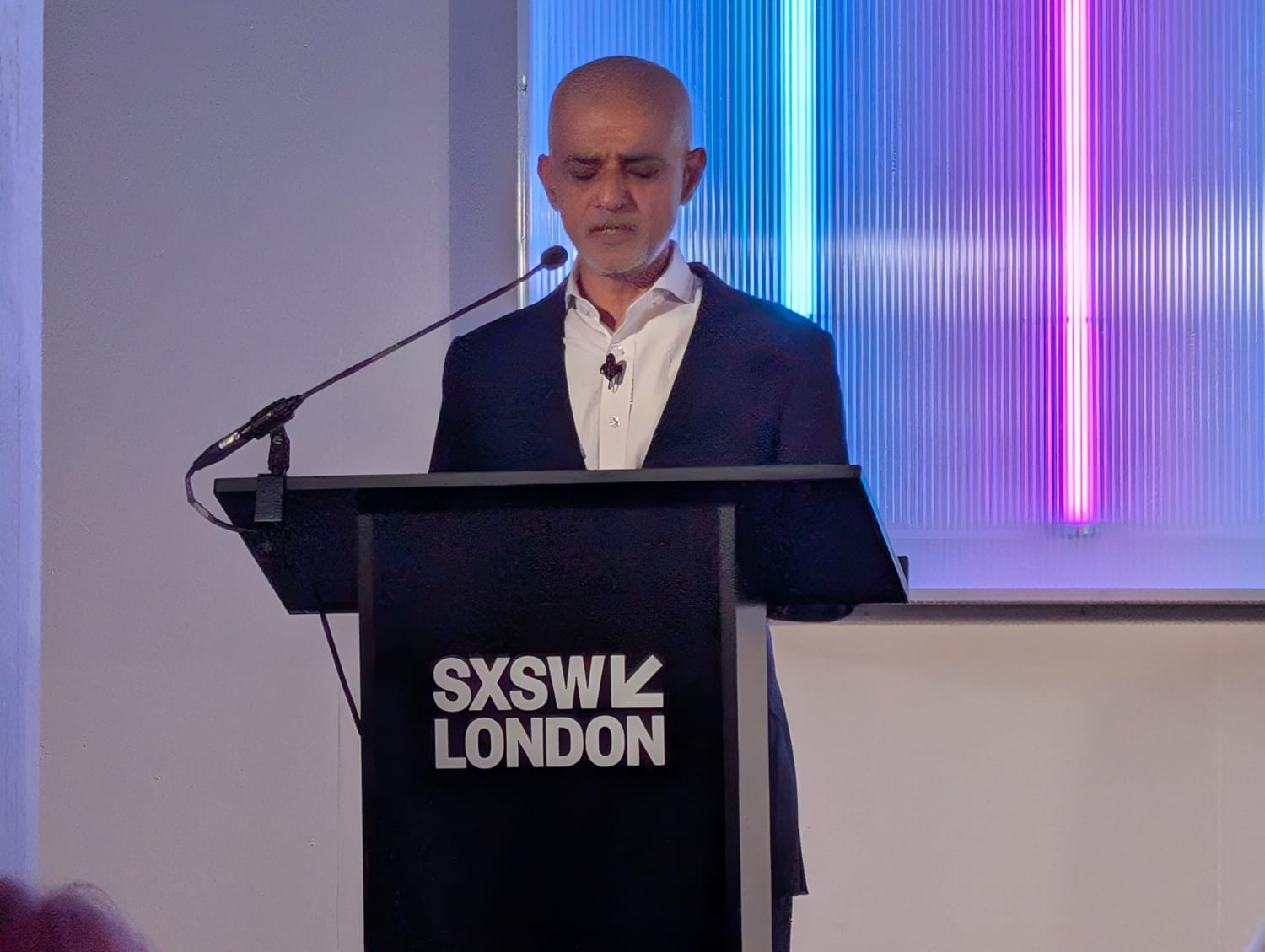 London mayor Sadiq Khan, an Asian man with shaved head in a navy suit speaks at a lectern in front of blue and pink neon strip lights.