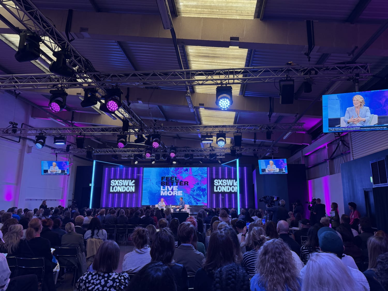 Two people on stage in the distance in front of a colourful backdrop. There are lots of lighting rigs across the ceiling and screens high up in the room. One features speaker Esther Perel, a small blond woman in a pale blue suit.