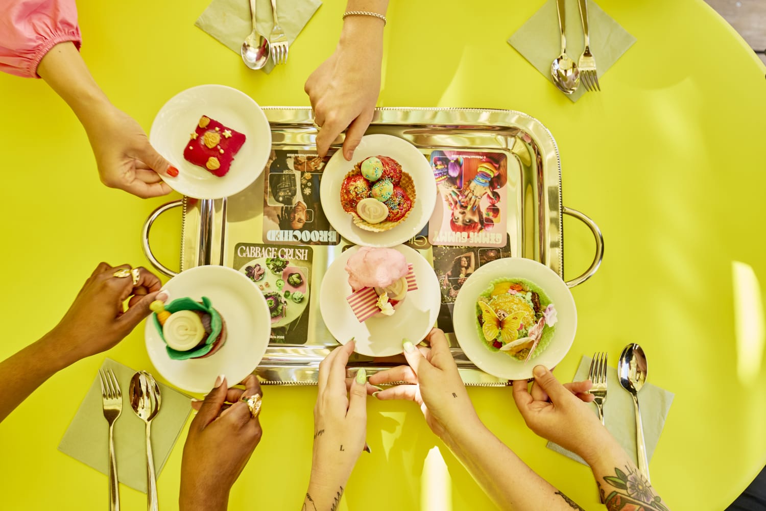 People participating in the patisserie experience at Pinterest beach at Cannes. A view of hands holding plates of cakes on a green table cloth covered with cutlery and napkins.