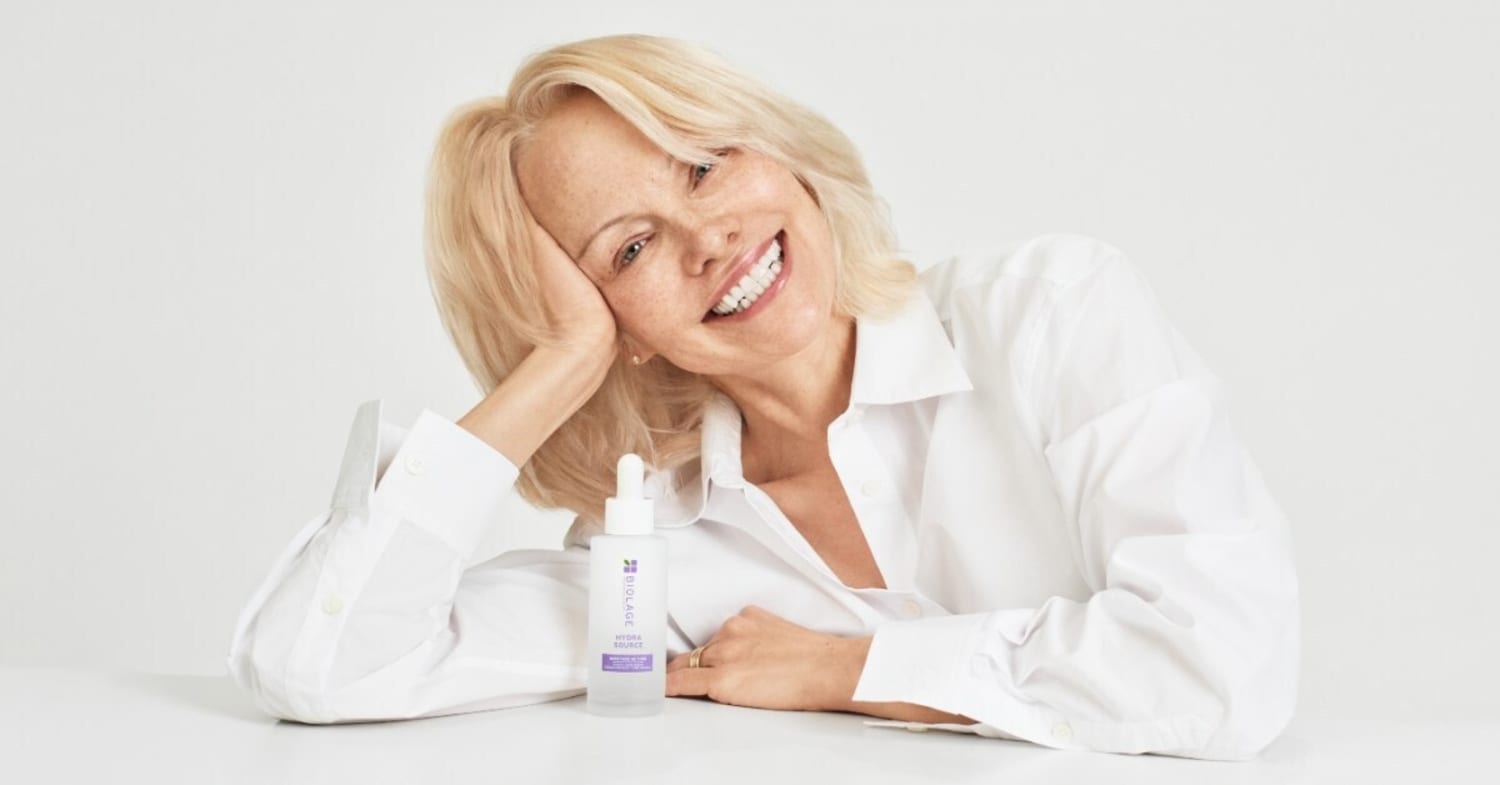Biolage x Pamela Anderson - a blond Pamela Anderson smiles at the camera, wearing a white shirt and leaning her head on her elbow.