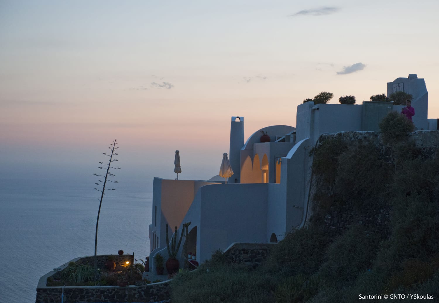 A scenic, dusk view of whitewashed buildings nestled on a cliffside in Santorini, Greece. Soft lights glow from within the architecture as the sun sets over the ocean, casting a peaceful, pastel-colored gradient across the sky.
