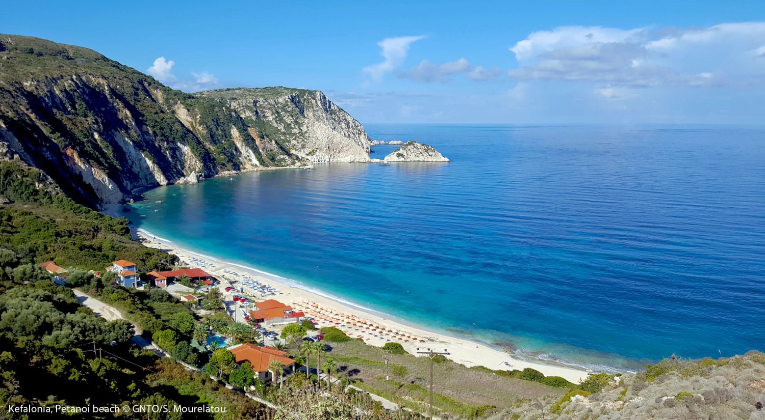 An elevated, panoramic view of Petani Beach in Cephalonia, Greece. The image showcases a sweeping white-sand crescent beach meeting calm, turquoise waters. Steep, green-covered cliffs line the coast, stretching out into the deep blue Ionian Sea under a clear sky.