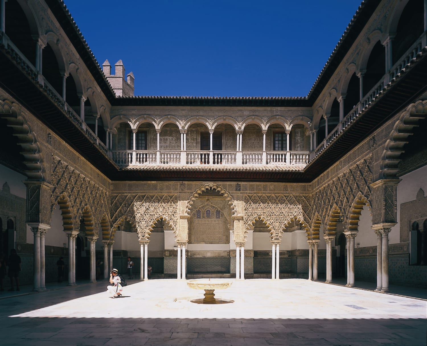 The sunny inner courtyard of the Alcázar of Seville, Spain, showing the Courtyard of the Maidens (Patio de las Doncellas). The courtyard features a white tiled floor, a central fountain, and is surrounded by stunning Mudéjar architecture with intricate plasterwork arches, columns, and an upper-level gallery under a bright blue sky.