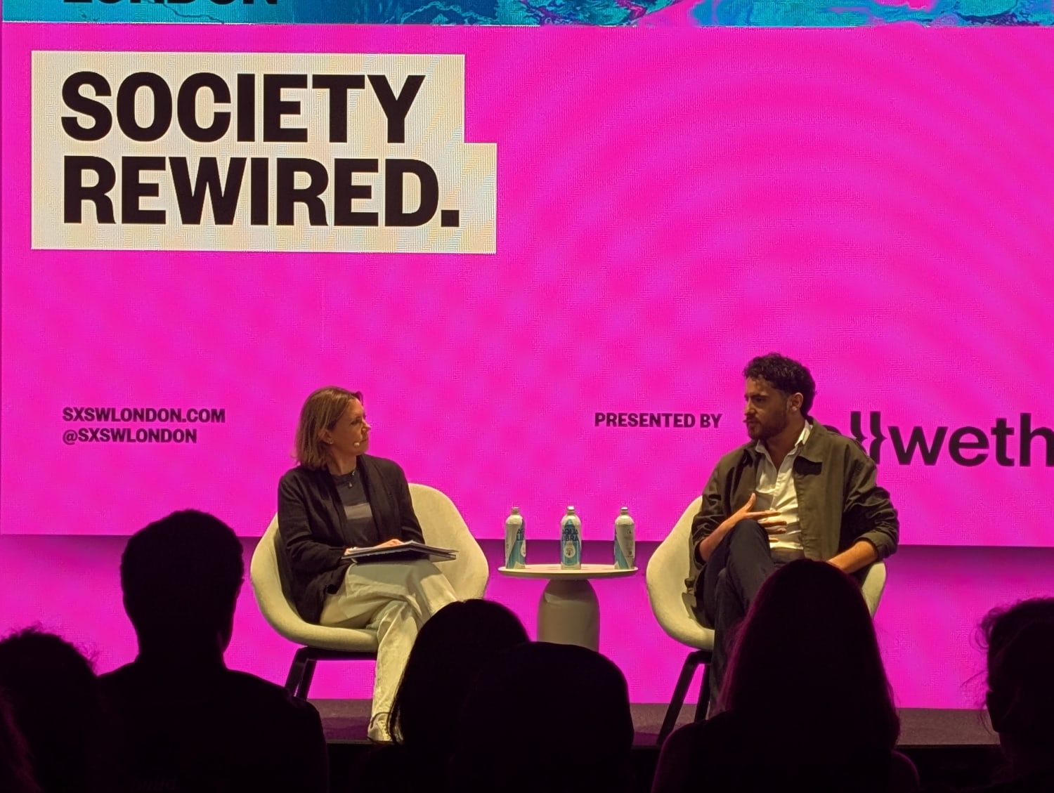 Man and woman on stage in front of a pink screen with the title Society Rewired.