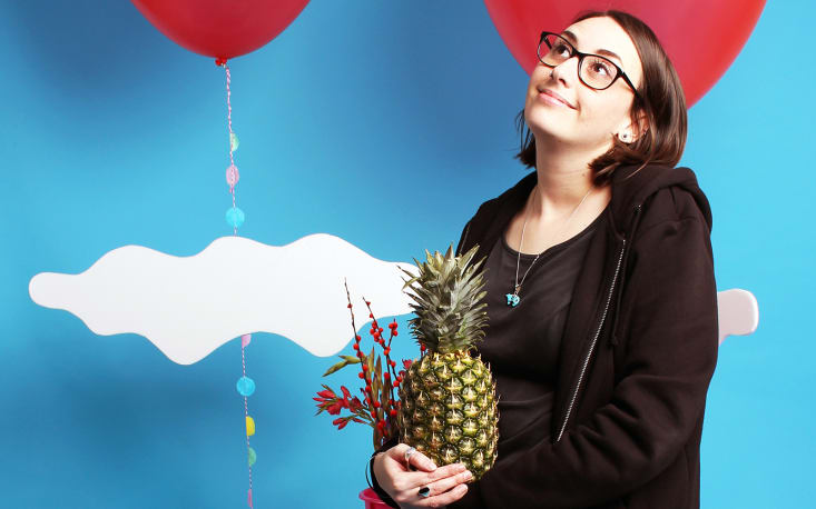 Woman in back tracksuit jacket holding a pineapple. Red balloons and clouds in back ground.