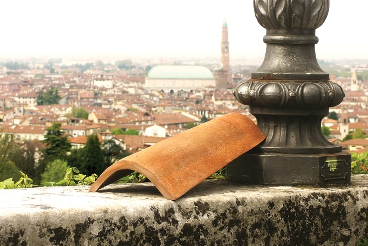A curved terracotta-colored tile rests on a stone ledge in front of a black lamppost. An Italian city lined with terracotta-tiled rooftops stretches out in the background.